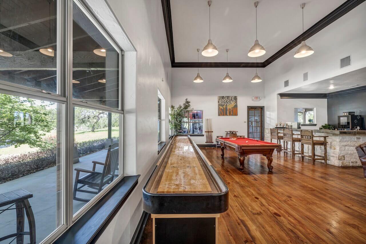 A spacious, well-lit game room featuring a shiny shuffleboard table, a pool table, and modern hanging lights, with a view of the outdoors through large windows.
